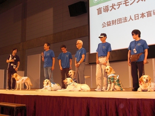 ステージ上でハーネスの会参加のみなさんと盲導犬が一列にならんでいる写真