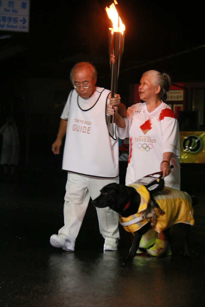 左手に盲導犬ヴォイスをつれ、右手で聖火を高くあげて走る岩谷さんの写真