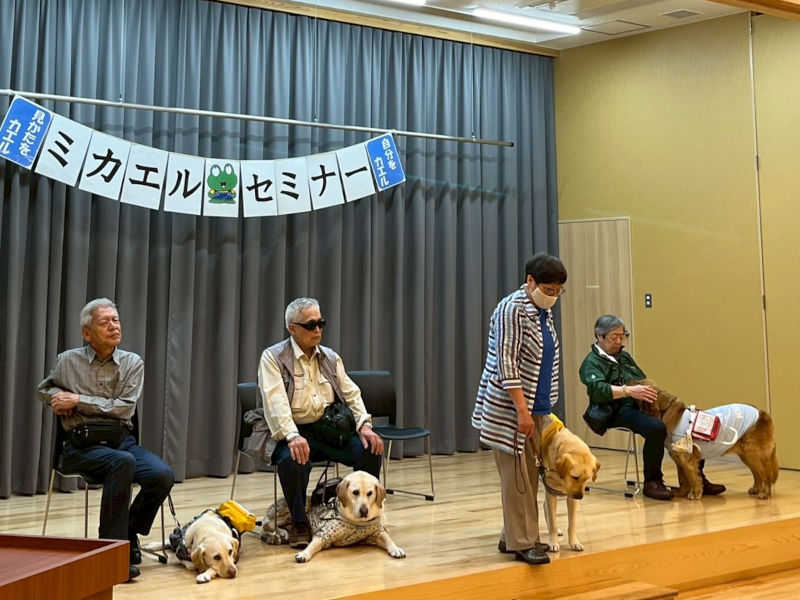 ミカエル・セミナー 盲導犬教室 ステージにユーザー4人と盲導犬4頭が並んでいる写真